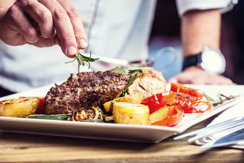 Chef In Hotel Or Restaurant Kitchen Cooking Only Hands. Prepared Beef Steak With Vegetable Decoration.